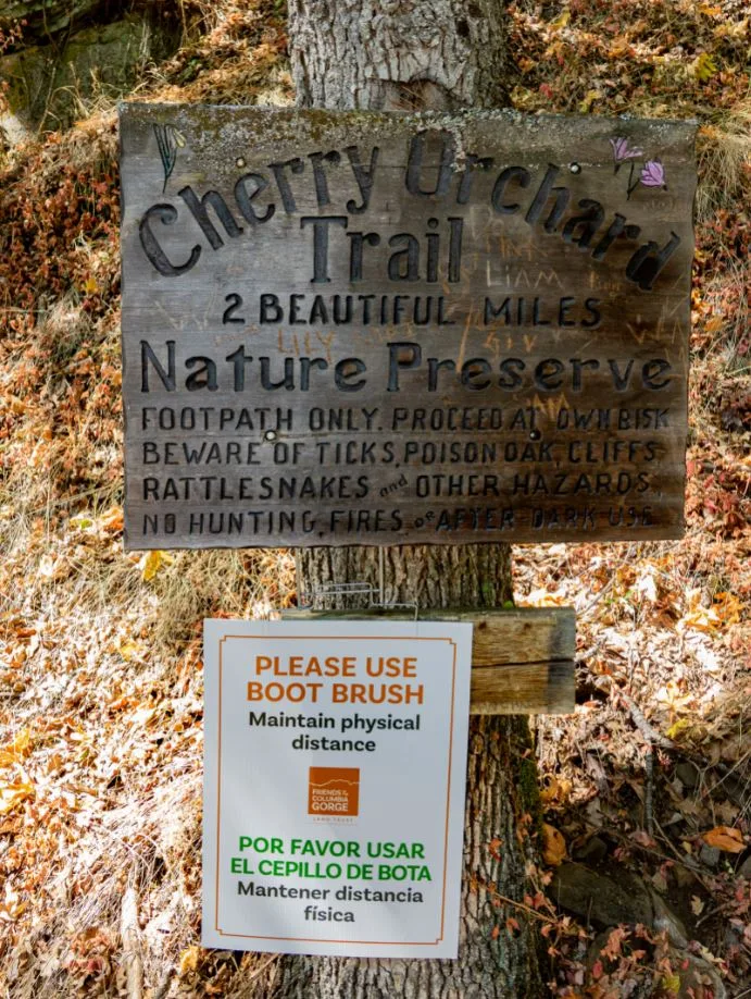 Photo: The original wood-carved Lyle Cherry Orchard trail sign is supplemented by a bilingual advisory sign. (Richard Kolbell)