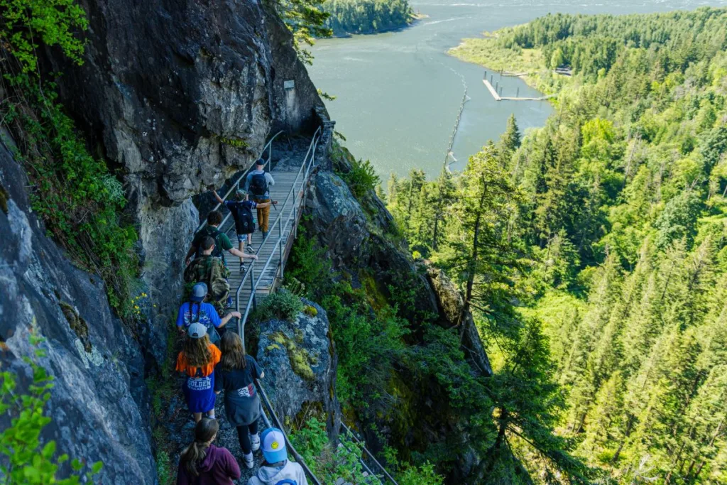 People hiking along a rock wall face on a rocky trail with a forest in the foreground and the columbia river in the background