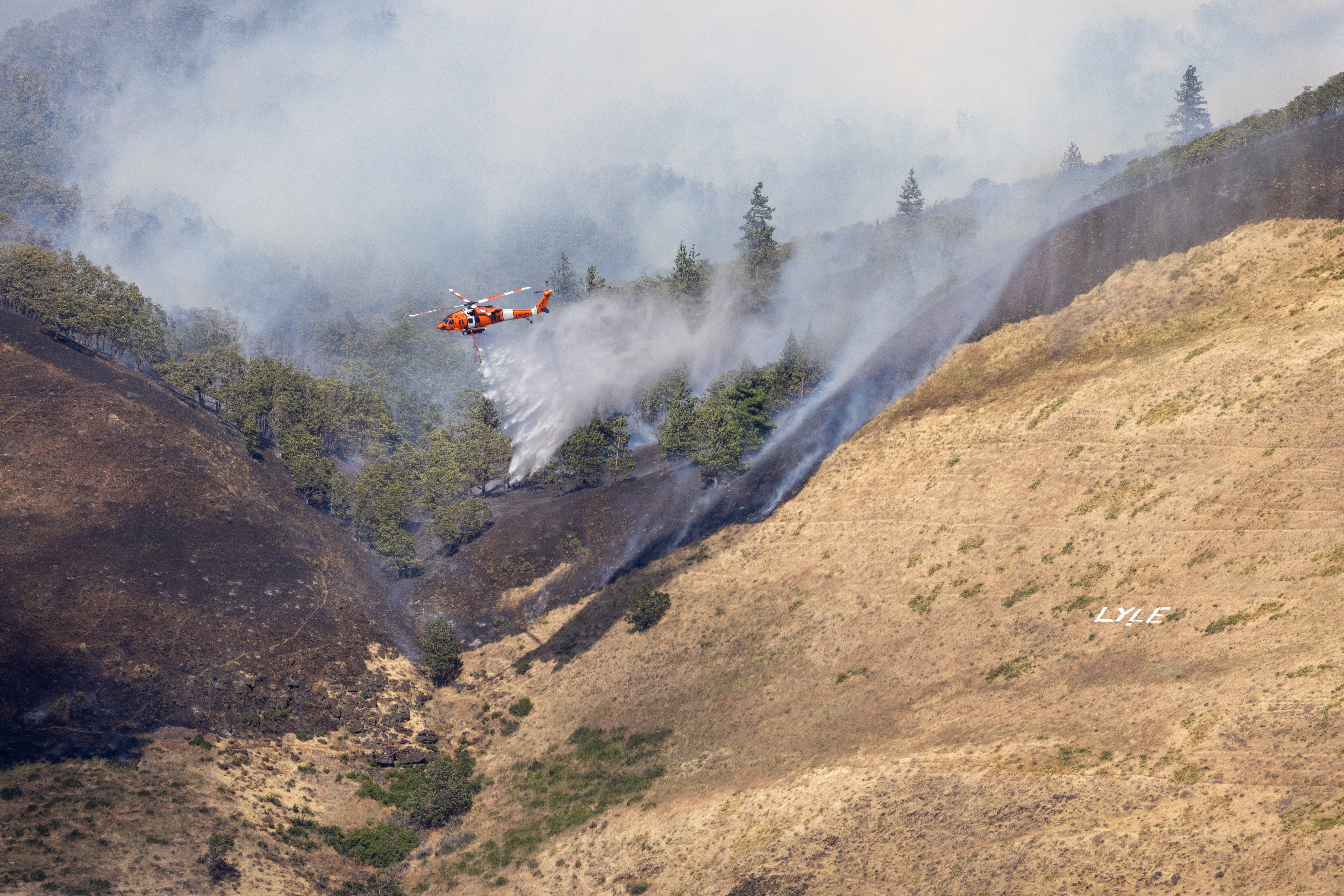 Wildfire burning through oak forest and grasslands as a helicopter drops water.